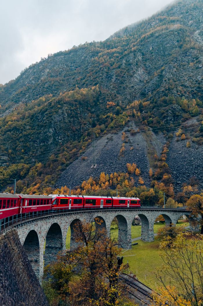 Red train traversing a scenic arch bridge in the Swiss Alps during autumn.